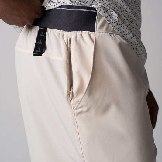 Gray background, close up image of the JOOLA logo tag on the side of a pair of Men's Club Shorts, sandstone color.