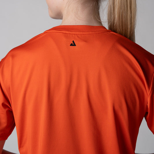 Gray background, a close up image of the black JOOLA trinity logo on the back of a female model wearing a JOOLA women's court crop tee, poppy red.
