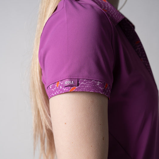 Gray background, close up on the raised JOOLA word mark logo in a JOOLA women's novelty polo dress, passion purple.