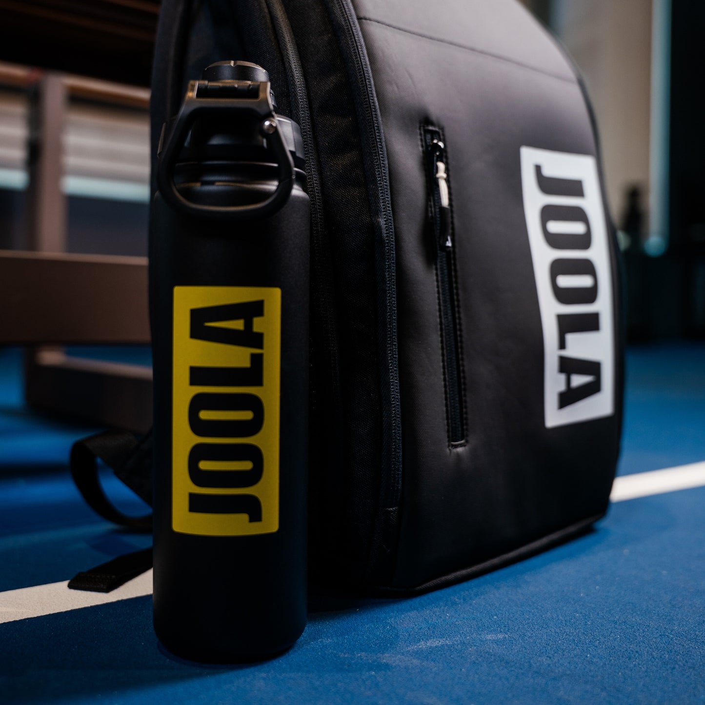 The black and yellow JOOLA water bottle positioned next to the JOOLA Vision Backapack sitting on an indoor pickleball court.