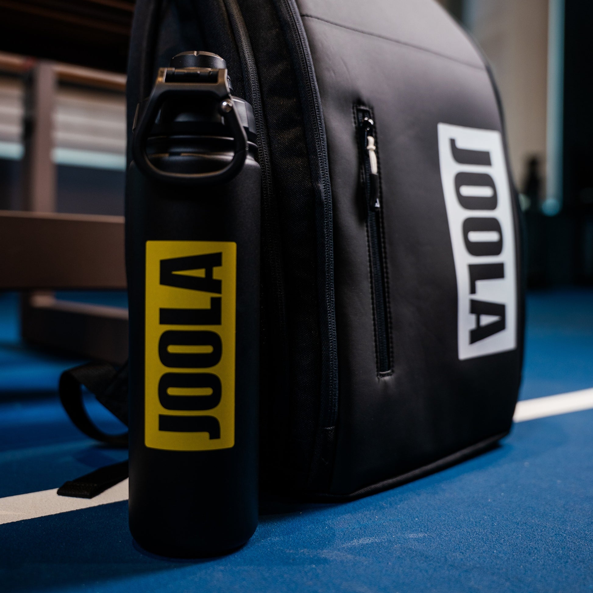 The black and yellow JOOLA water bottle positioned next to the JOOLA Vision Backapack sitting on an indoor pickleball court.