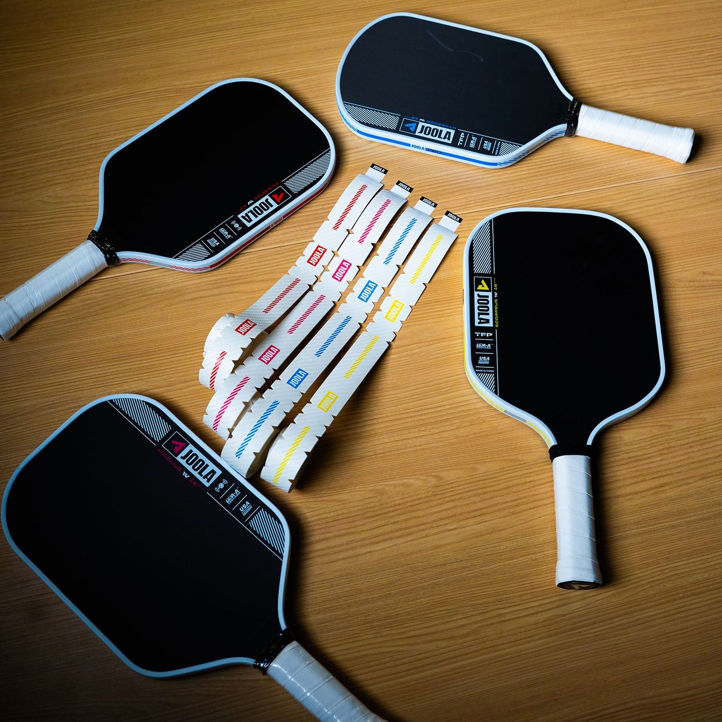 JOOLA Pro IV Pickleball paddles laying on a wood background with four un-rolled JOOLA Pro IV Edge Guard tape colors in red, pink, blue and yellow in between the four paddles.
