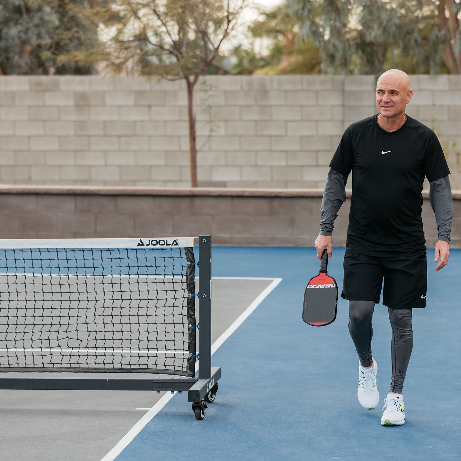 Image of Andre Agassi with JOOLA paddle in hand walking on a pickleball court next to a JOOLA pro pickleball net.